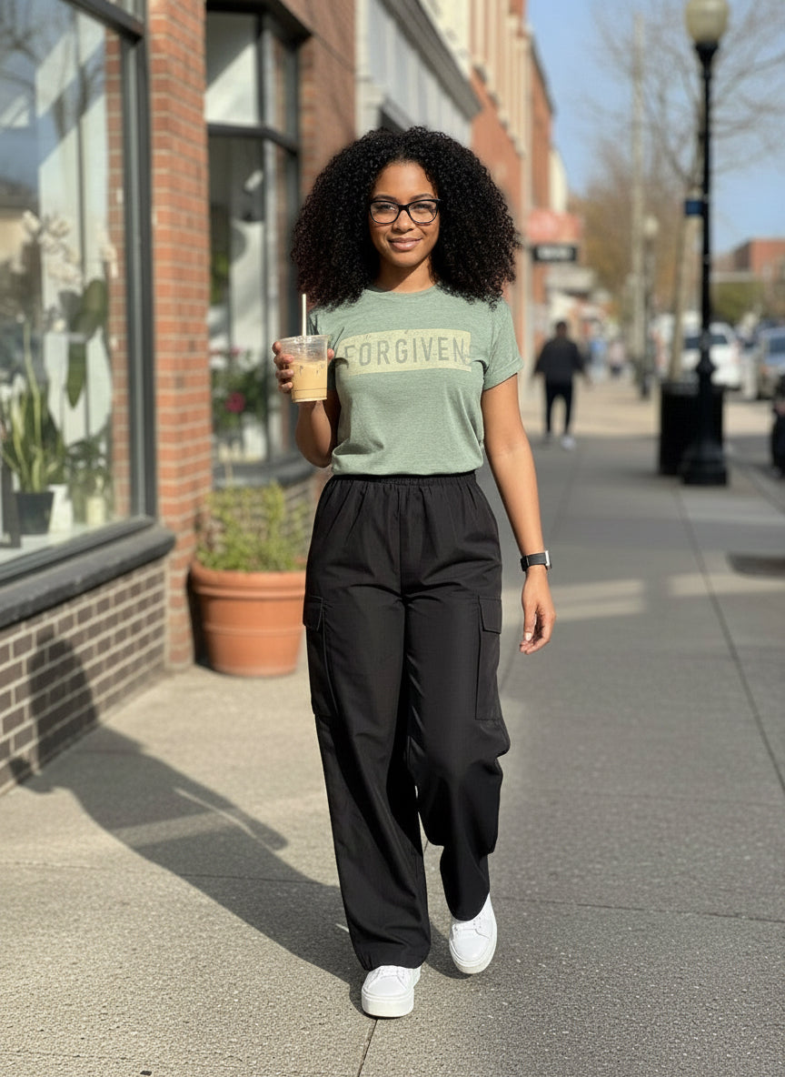Woman walking on a sidewalk holding a drink, wearing a green t-shirt and black pants.