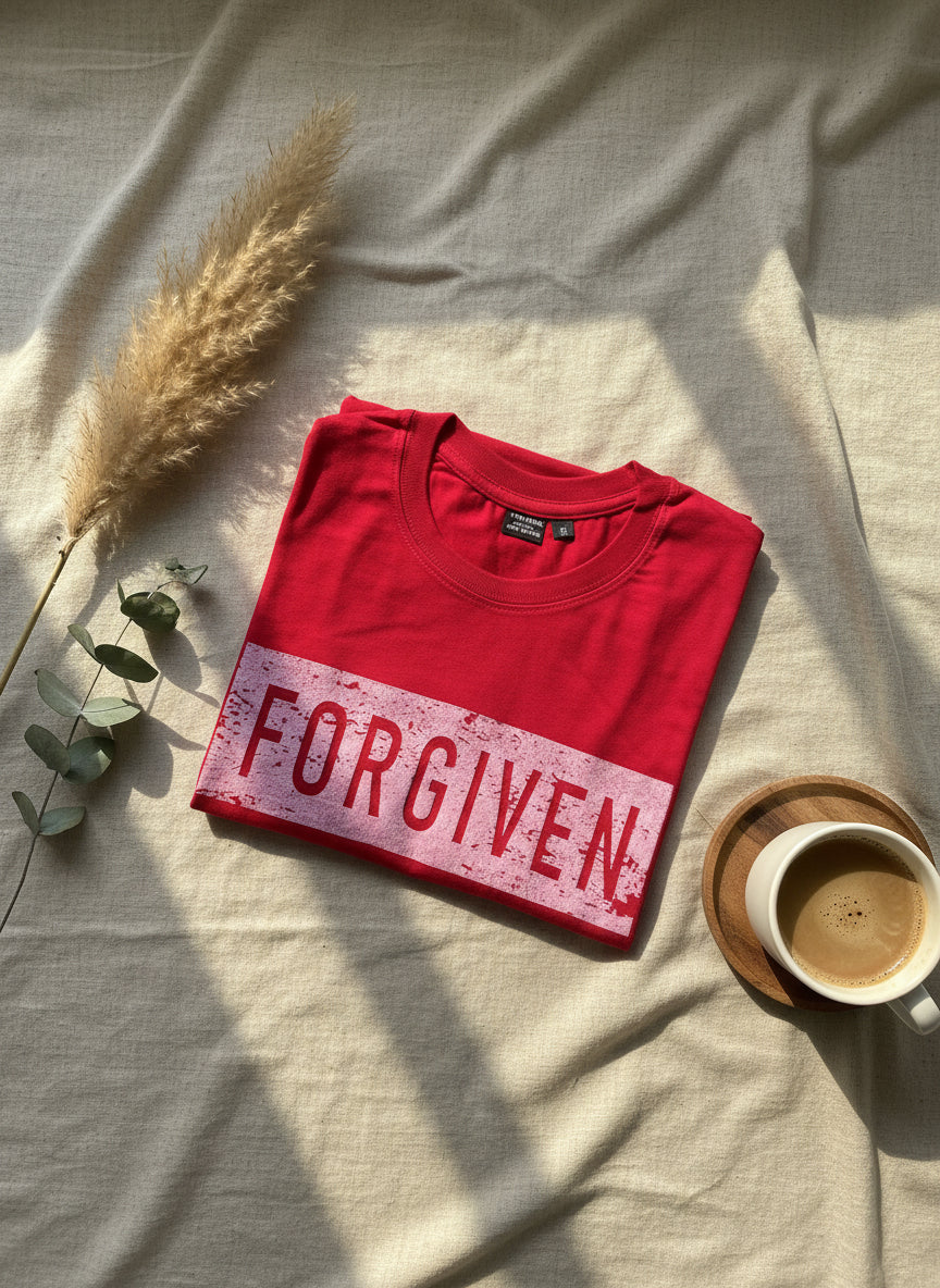 Red t-shirt with 'FORGIVEN' text on a light fabric background with a cup of coffee and dried plants.