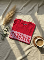 Red t-shirt with 'FORGIVEN' text on a light fabric background with a cup of coffee and dried plants.