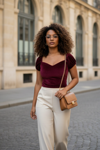 Woman in burgundy top and white pants walking on a street with a beige building in the background