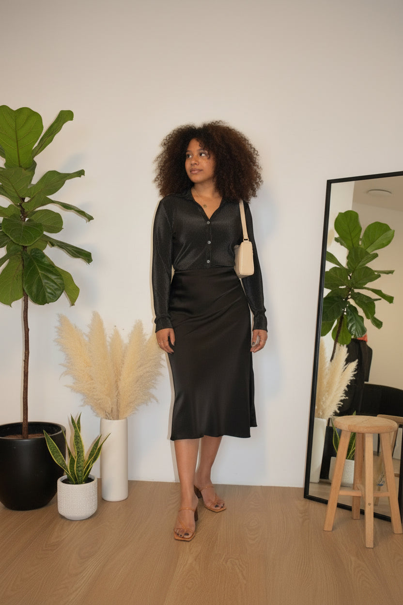 Woman in a black satin dress standing in a room with plants and a mirror.