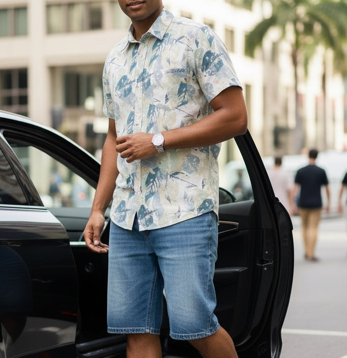 Man in a floral shirt and denim shorts getting out of a car on a city street.