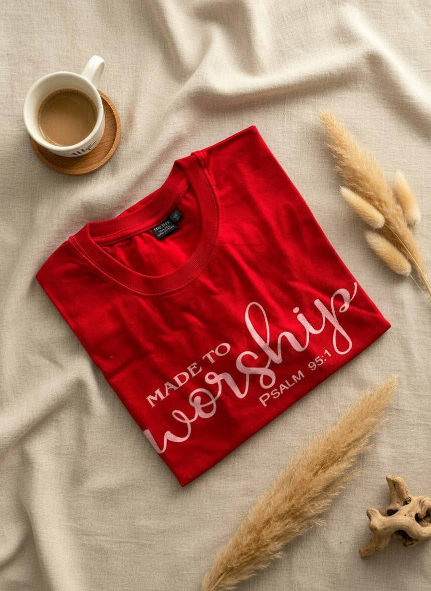Red t-shirt with 'Made to Worship' text on a beige surface with a cup and dried plants.