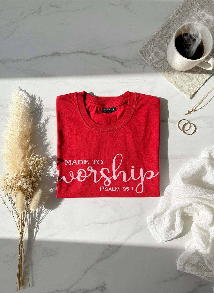 Red t-shirt with 'Made to Worship' text on a marble surface with a cup of coffee and dried plants.