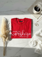 Red t-shirt with 'Made to Worship' text on a marble surface with a cup of coffee and dried plants.