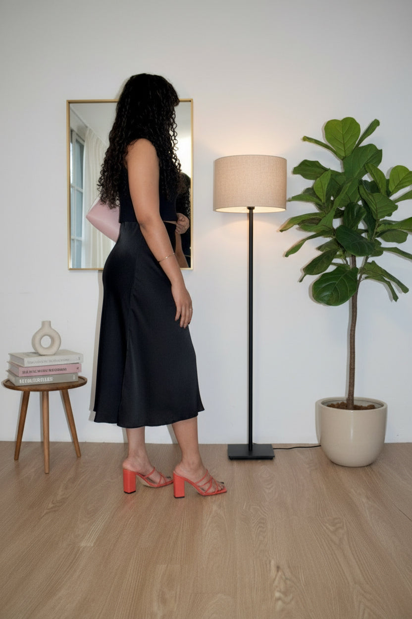 Woman in a black dress standing in a room with a lamp, mirror, and plant.