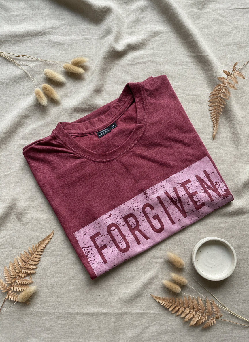 Folded maroon t-shirt with 'FORGIVEN' text on a light surface with decorative elements.
