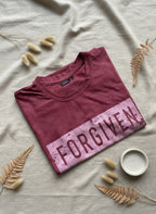 Folded maroon t-shirt with 'FORGIVEN' text on a light surface with decorative elements.