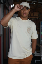 Person wearing a beige t-shirt with a logo and brown pants, standing indoors.