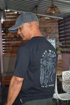 Man wearing a black t-shirt with a logo and text, bending over in an outdoor setting.
