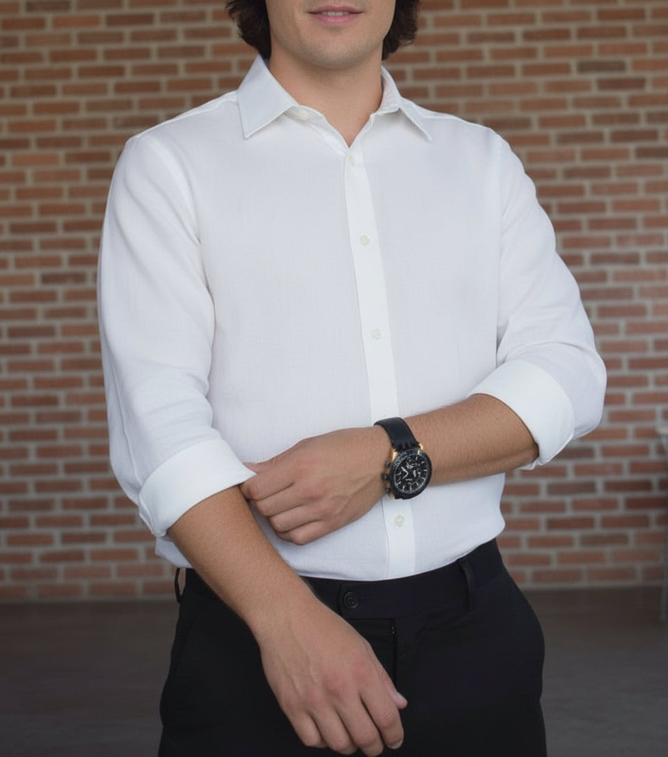 Man wearing a beige shirt and khaki pants with a watch, standing against a brick wall.